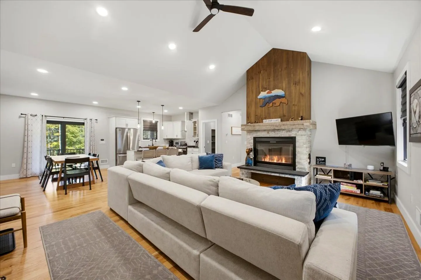Open-concept living room and kitchen featuring a stone fireplace with a wood-paneled accent, a large sectional couch, and a dining area.
