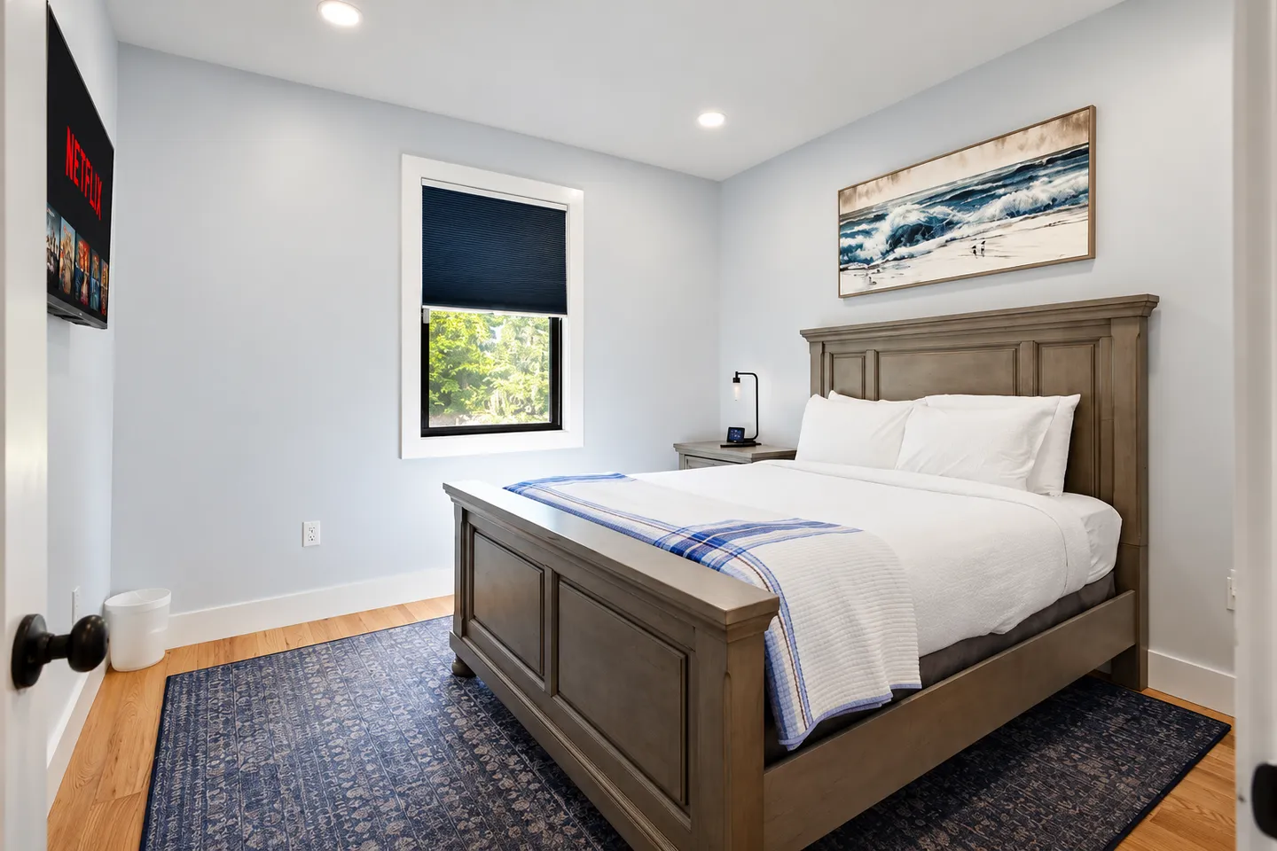Bedroom with a wood-framed bed, white bedding, patterned blue rug, wall-mounted TV, and a window with a dark roller shade.