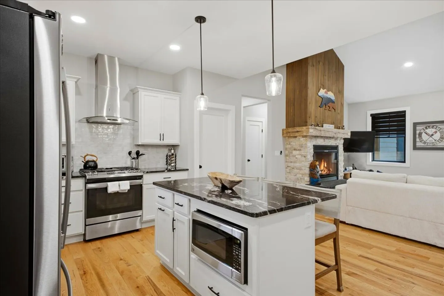 Open-concept kitchen with white cabinets, dark island countertop, and a view into the living room featuring a stone fireplace.