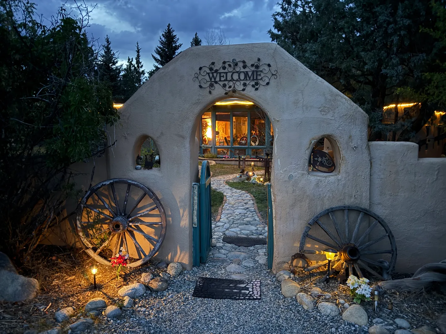 Stucco archway with a Welcome sign and open teal gate leading along a stone pathway toward a warmly lit home at dusk.