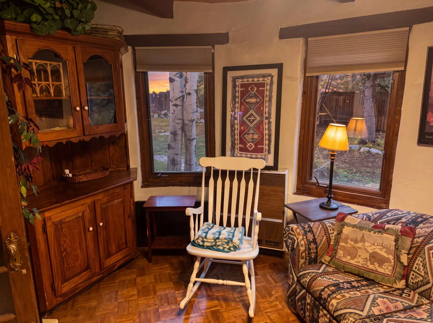 A cozy room corner featuring parquet flooring, a white wooden rocking chair, a patterned armchair with a bear accent pillow, a corner wooden hutch, and windows framing outdoor trees.