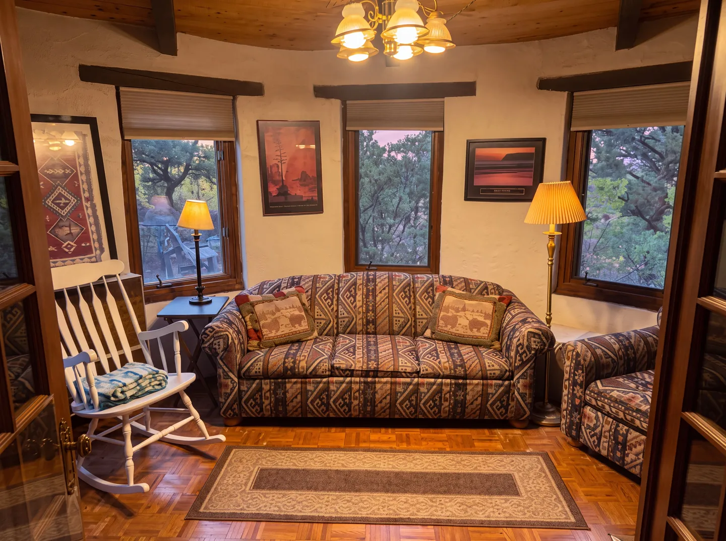A rounded sitting room featuring patterned Southwestern-style sofas, a white rocking chair, and multiple windows looking out toward trees.