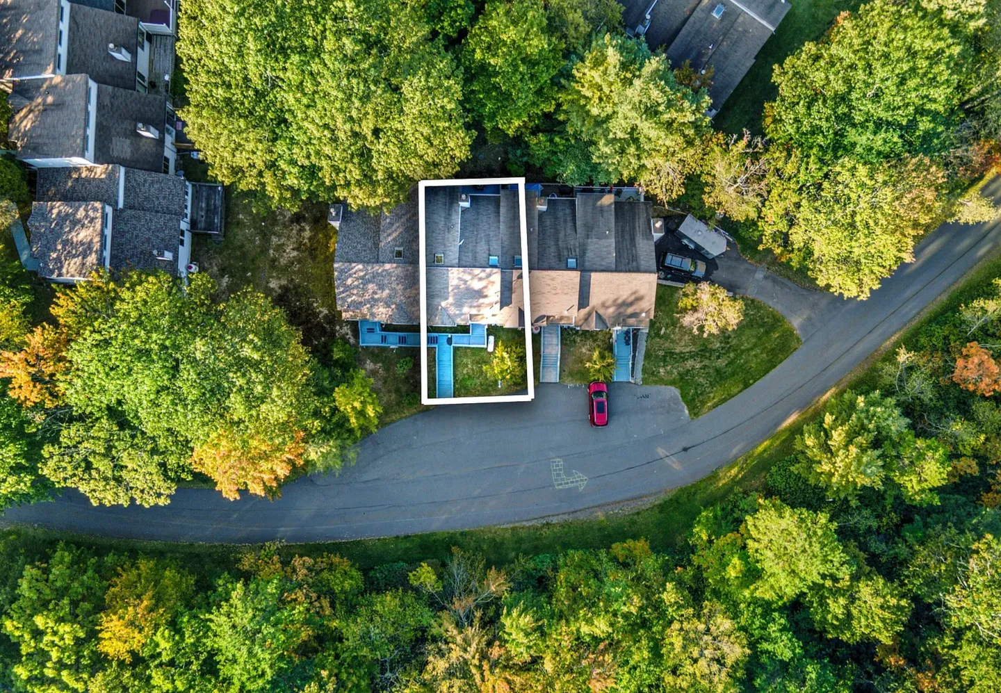 Overhead aerial view of a townhome unit outlined in white, nestled among dense green trees with a curved paved road and driveway in front.