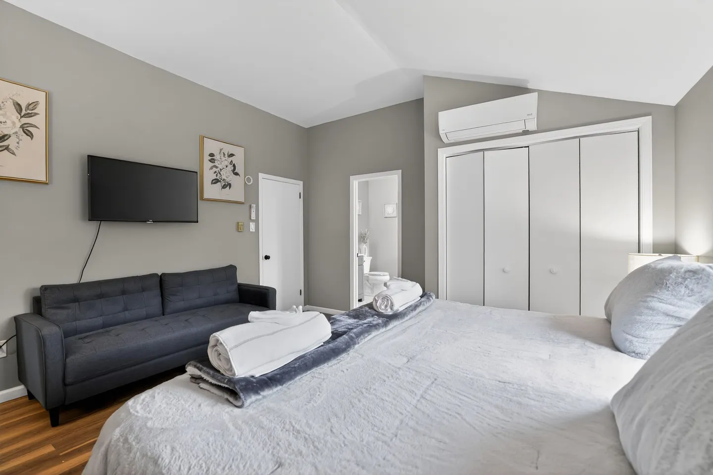 Bedroom with a bed in the foreground, a dark gray sofa beneath a wall-mounted TV, white closet doors, and an open doorway leading to a bathroom.