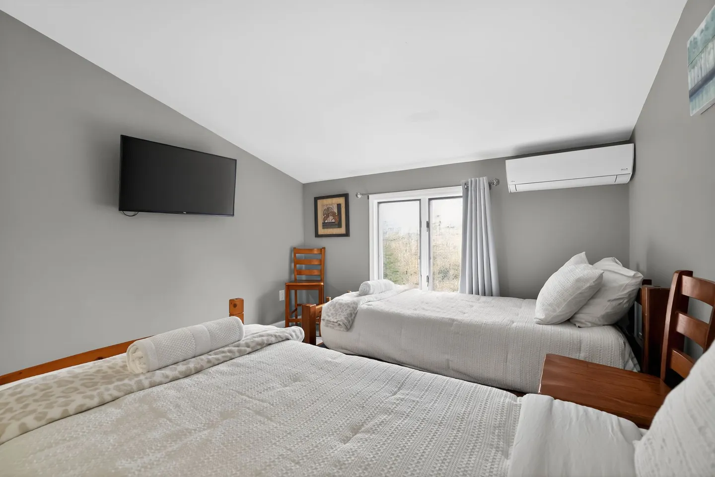 Bedroom with two beds made with white bedding, a wall-mounted TV, and a window in the background.