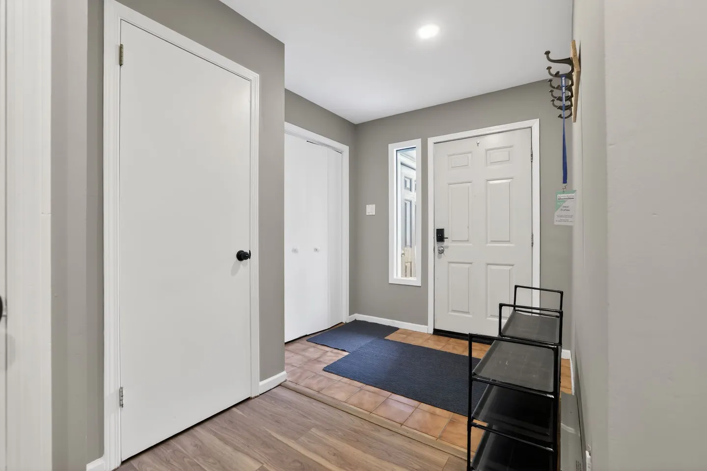 Front entry foyer featuring a white paneled front door, sidelight window, tiled landing with floor mats, and a shoe rack.