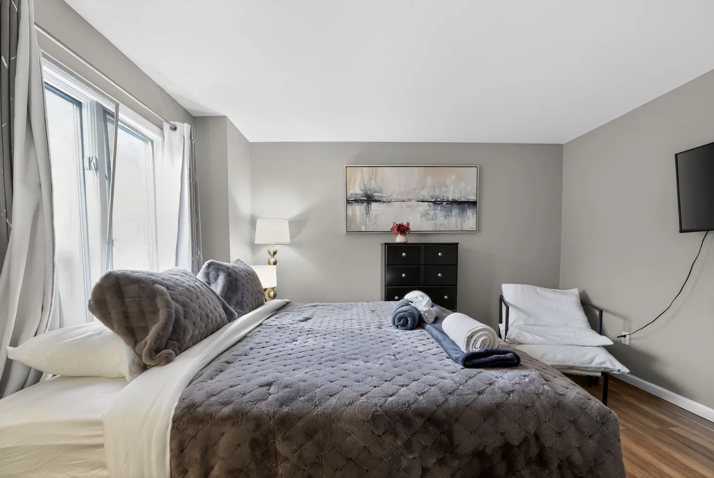 Bedroom with a large bed dressed in grey plush bedding, a black dresser, wall art, and a chair.