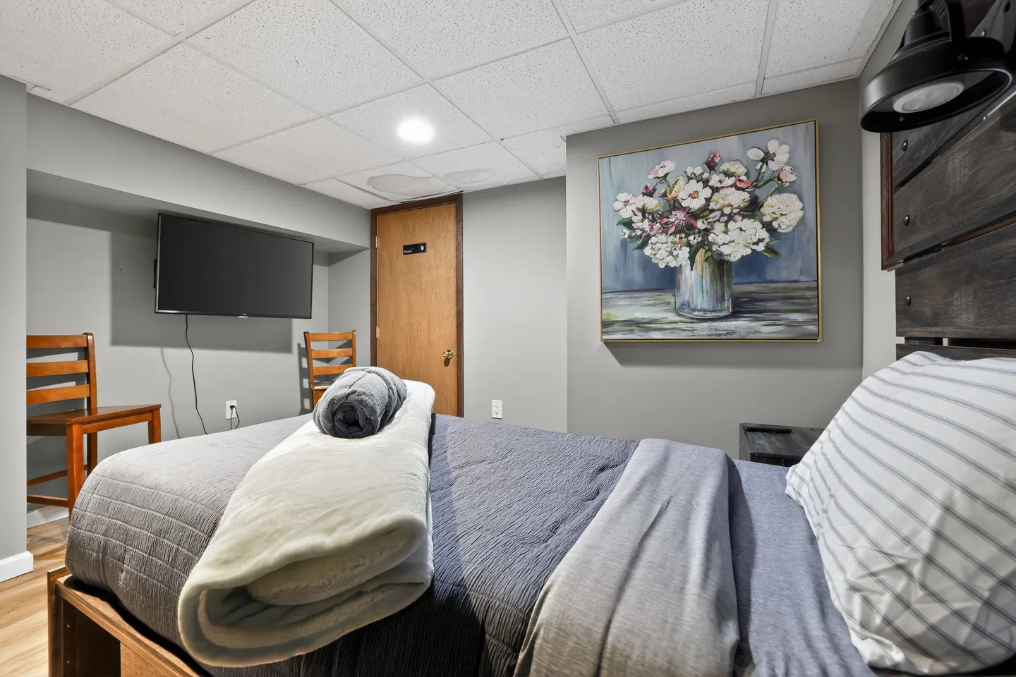 Bedroom featuring a made bed, wall-mounted television, floral artwork, and a drop ceiling.