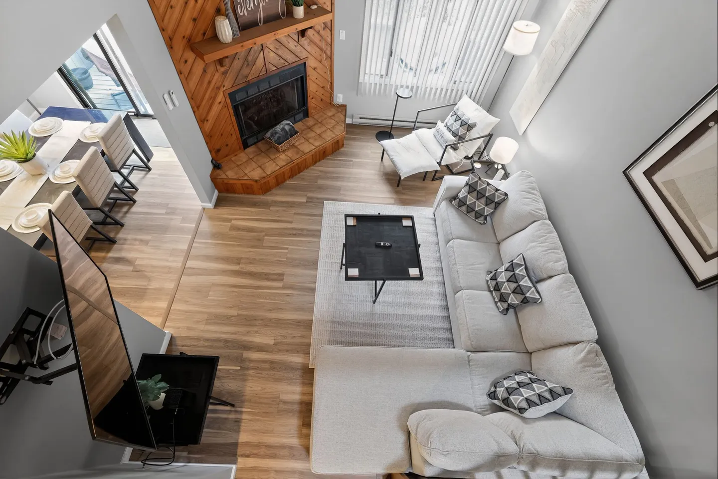 High-angle view of a living room with a wood-paneled fireplace, sectional couch, coffee table, and an adjacent dining area.