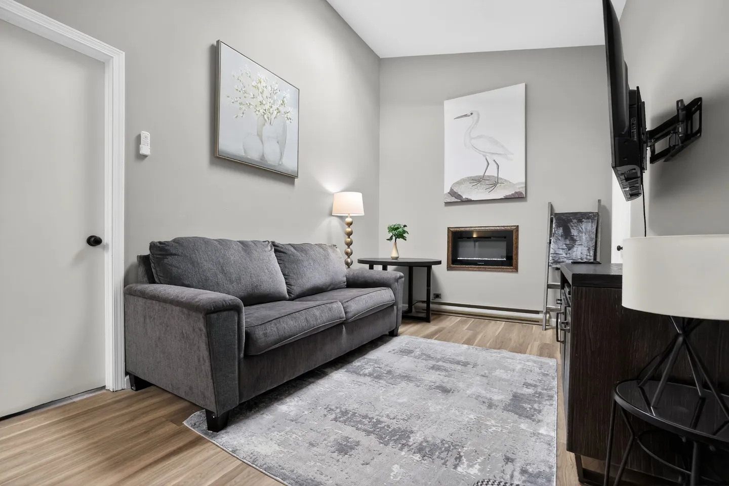 A living room area featuring a gray sofa, area rug, wall-mounted electric fireplace, and framed artwork.