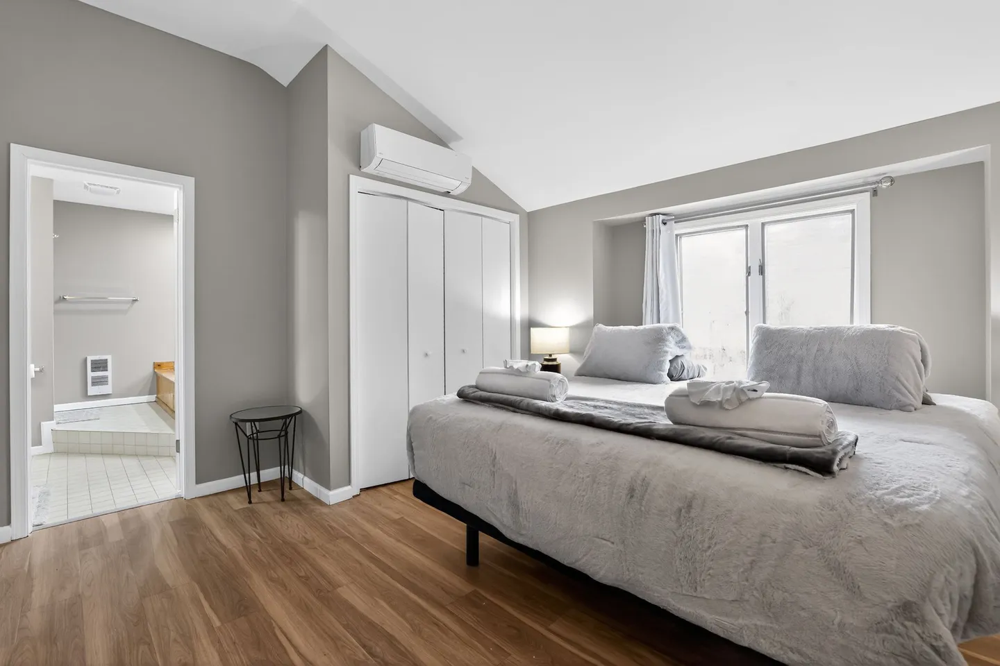Bedroom with a large bed dressed in grey linens, wood-style flooring, a mini-split AC unit, and an ensuite bathroom doorway.