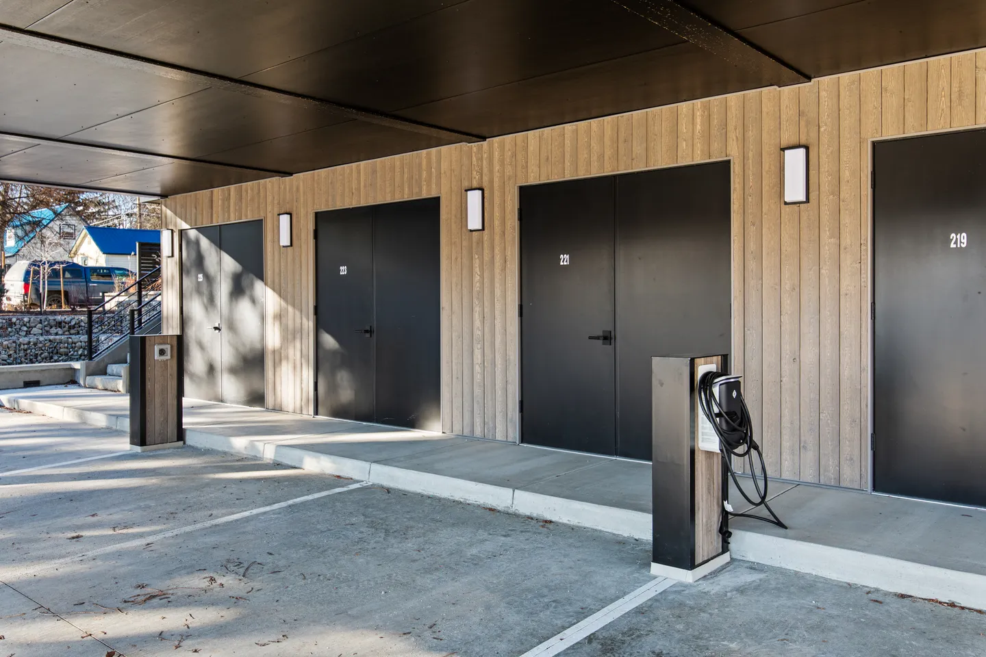 Covered parking area with concrete spaces, electric vehicle charging stations, and labeled black doors along wood-paneled walls.