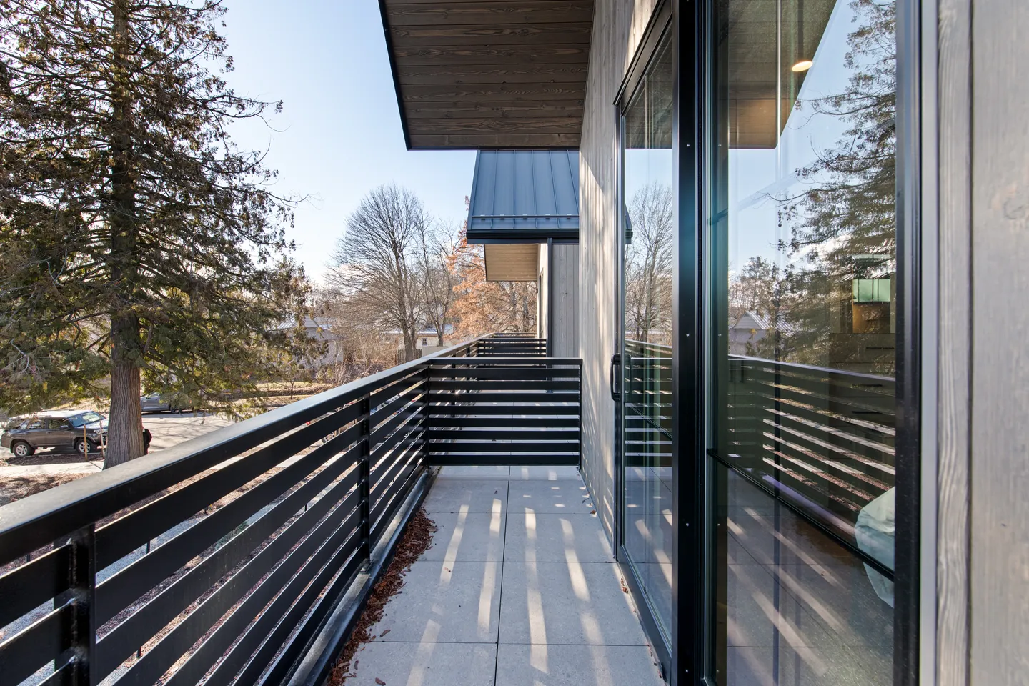 Outdoor balcony with dark horizontal metal railings, concrete pavers, sliding glass doors, and views of surrounding trees and blue sky.