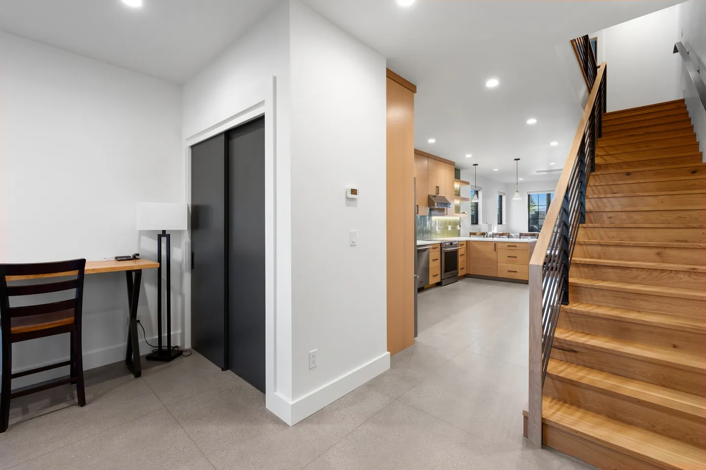 Hallway featuring a wooden desk and chair, dark sliding doors, wooden staircase, and a view into the open kitchen.