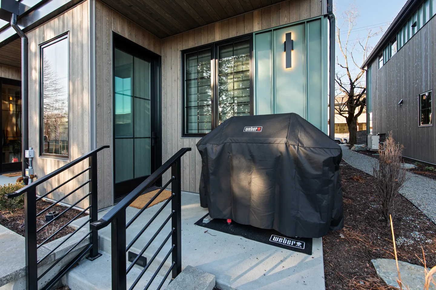 A covered outdoor barbecue grill sits on a concrete patio next to the entrance of a modern home with wood and teal siding.