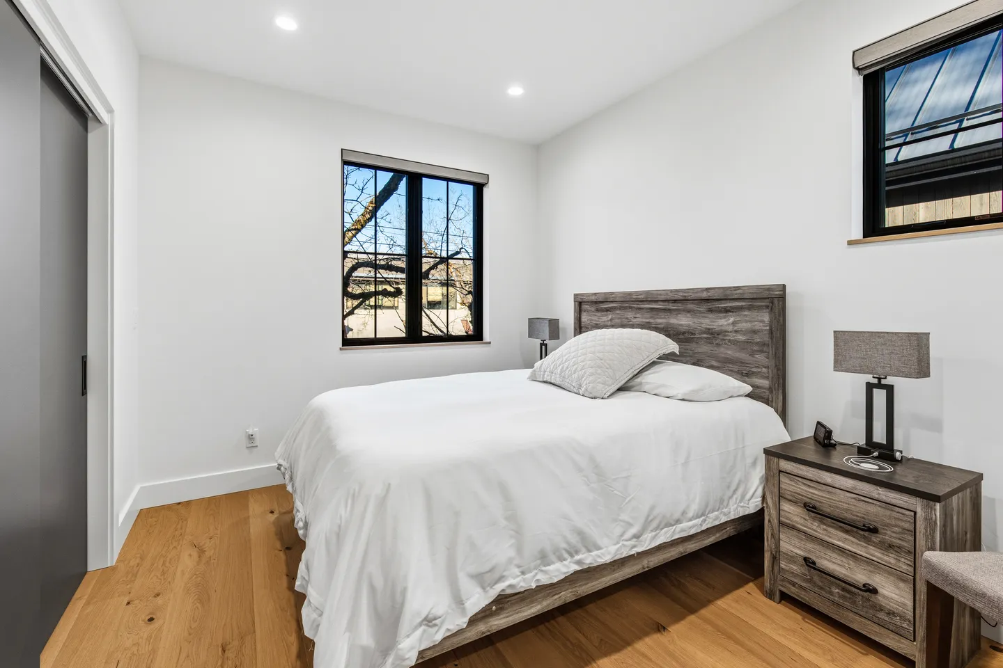 Bedroom with a rustic wood-look headboard, white bedding, wood flooring, black-framed windows, and a matching nightstand.