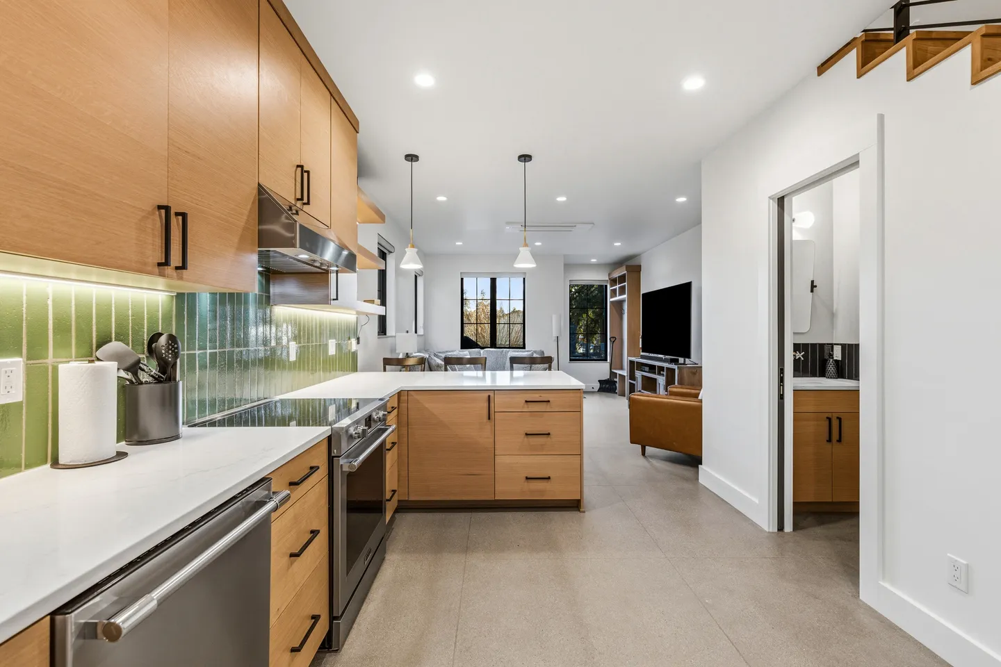 Open-concept modern kitchen with light wood cabinetry, green tile backsplash, and stainless steel appliances looking into the living area.