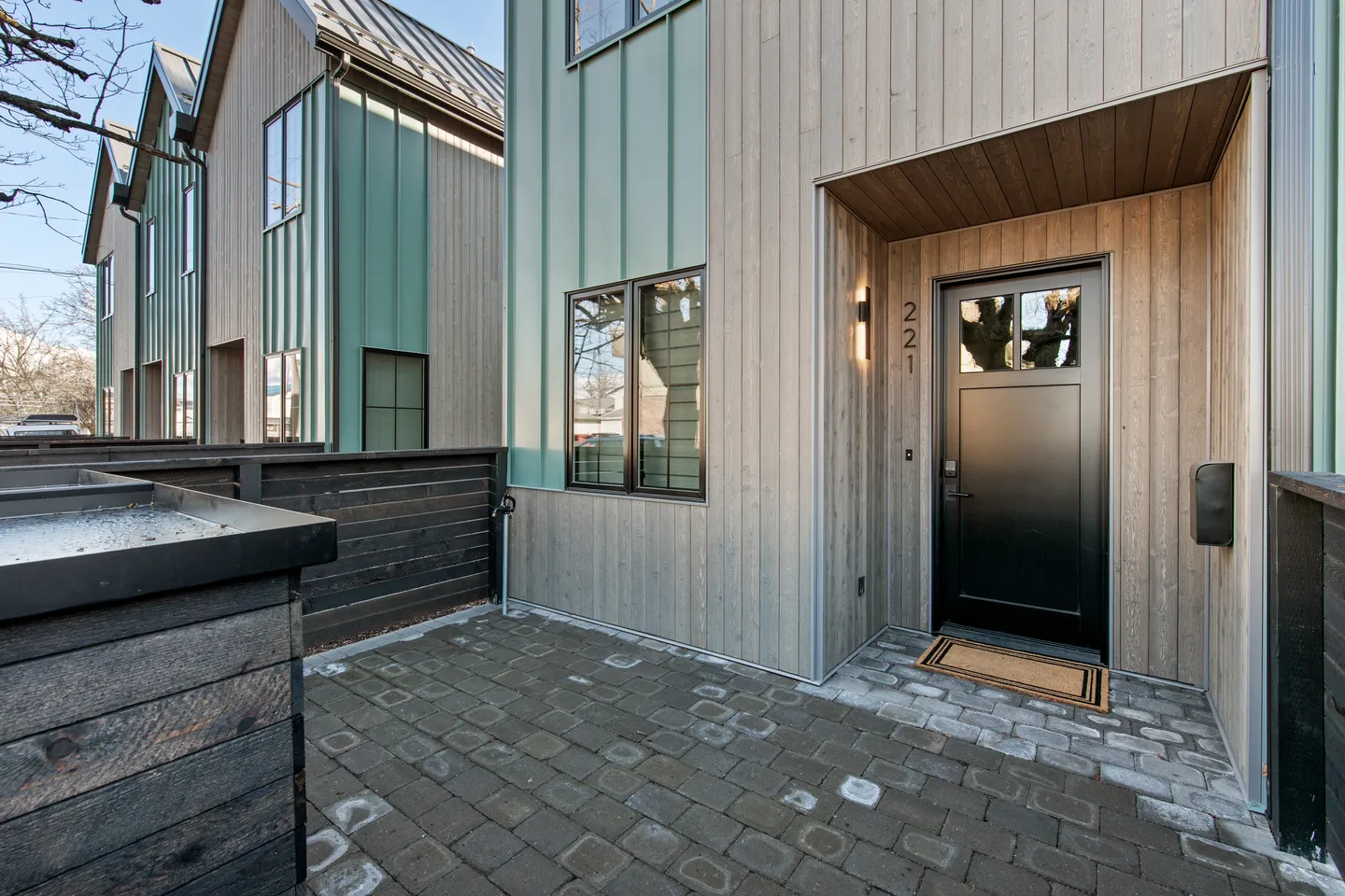 Exterior entryway of a modern home featuring vertical wood and teal siding, a black front door, and a brick paver patio.