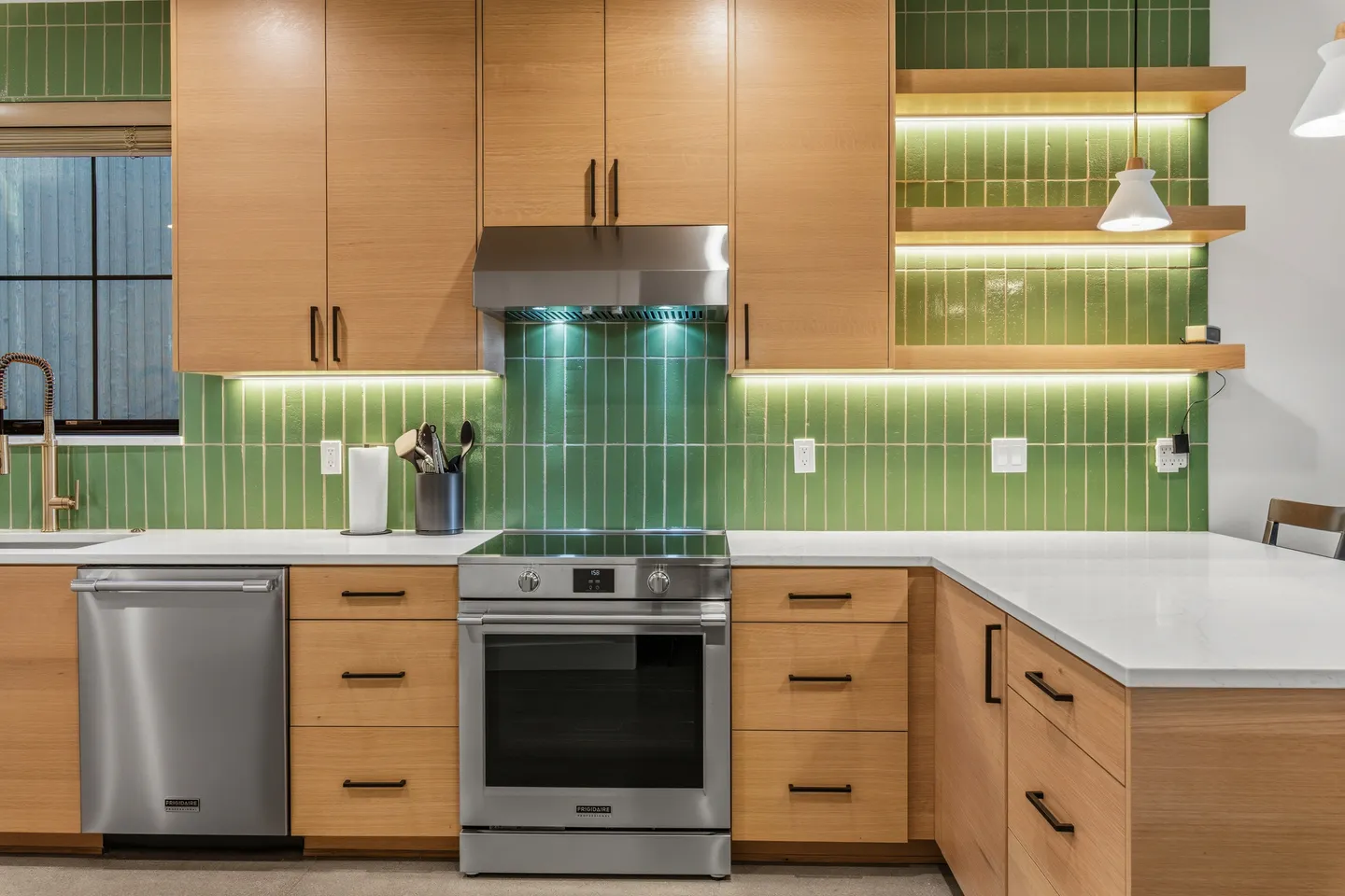 Modern kitchen with light wood cabinets, green vertical subway tile backsplash, white countertops, and stainless steel appliances.
