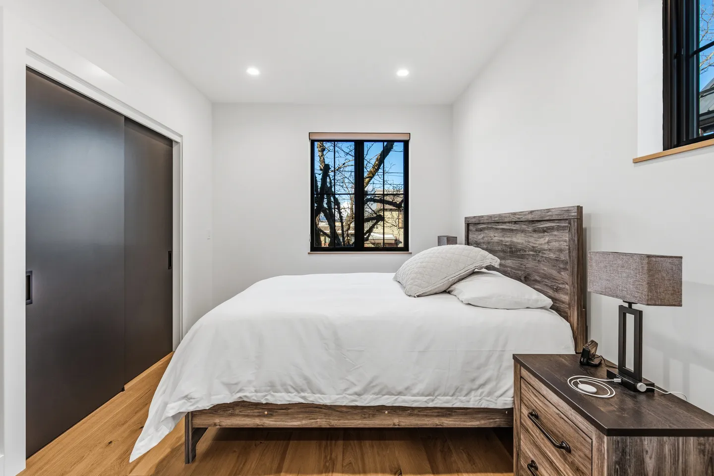Bedroom with a wooden bed frame, white bedding, hardwood floors, a nightstand, and a black-framed window.