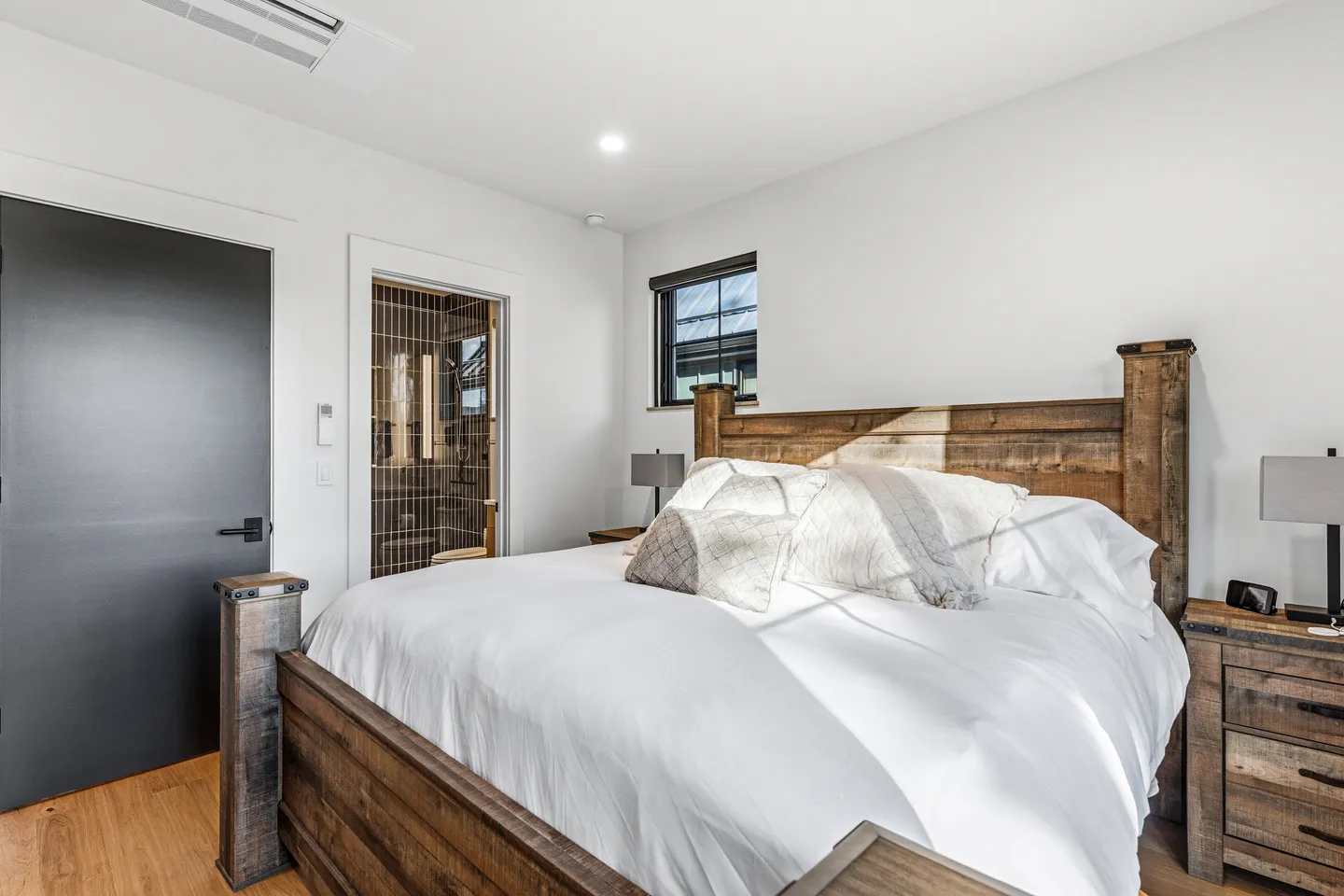 Bedroom with a rustic wood bed frame, white bedding, nightstands, and a doorway opening to an en-suite bathroom with dark tiled walls.