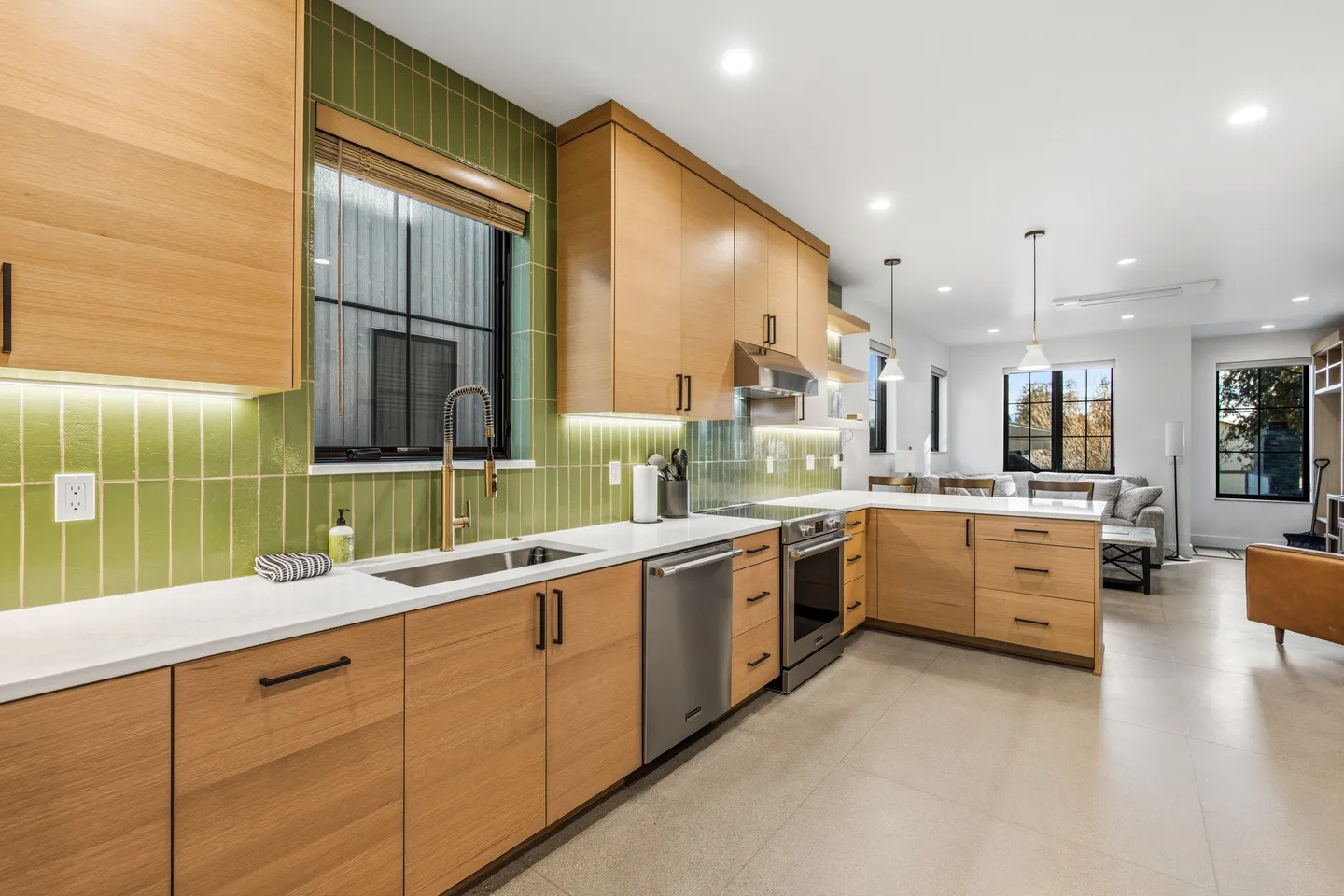 Kitchen with light wood cabinetry, green vertical tile backsplash, white countertops, and stainless steel appliances opening to a living area.