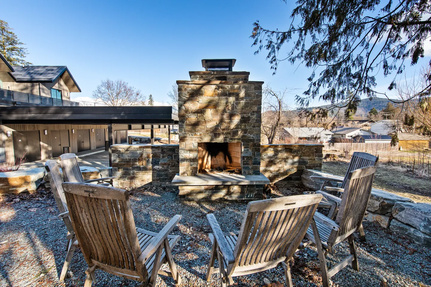 Outdoor stone fireplace with wooden armchairs on gravel under a clear sky.