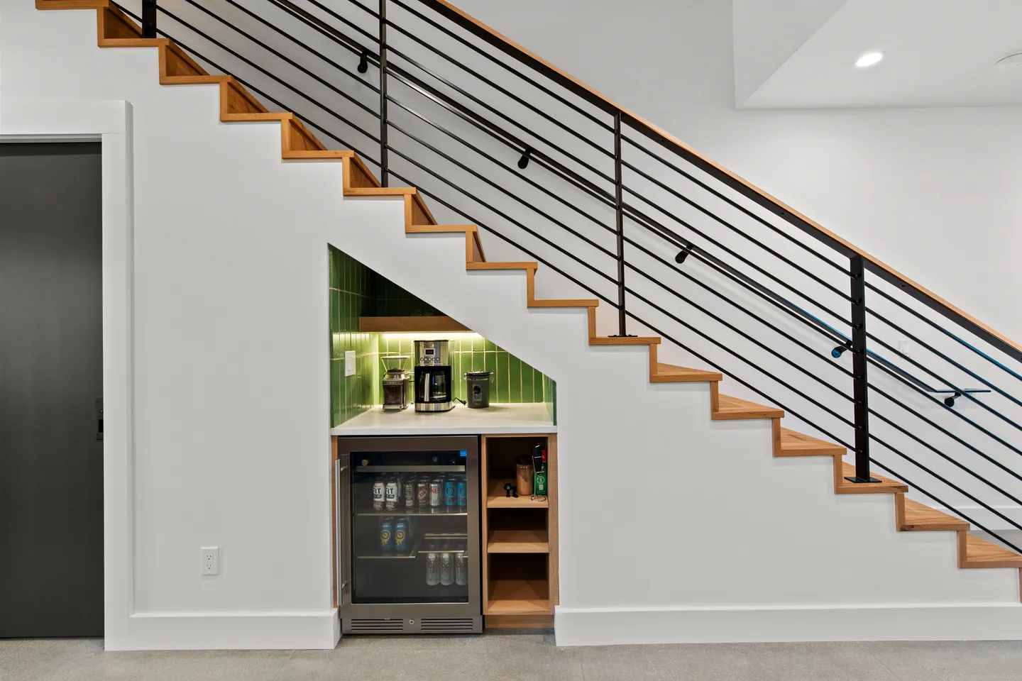 Beverage bar and mini fridge built into the nook beneath an indoor staircase with black metal railings.