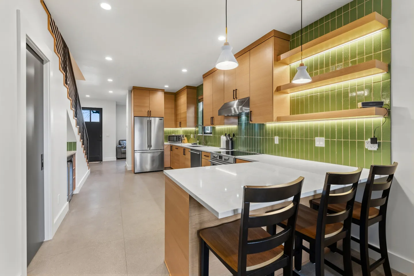 A modern kitchen with light wood cabinetry, green vertical tile backsplash, stainless steel appliances, and a peninsula with bar seating.