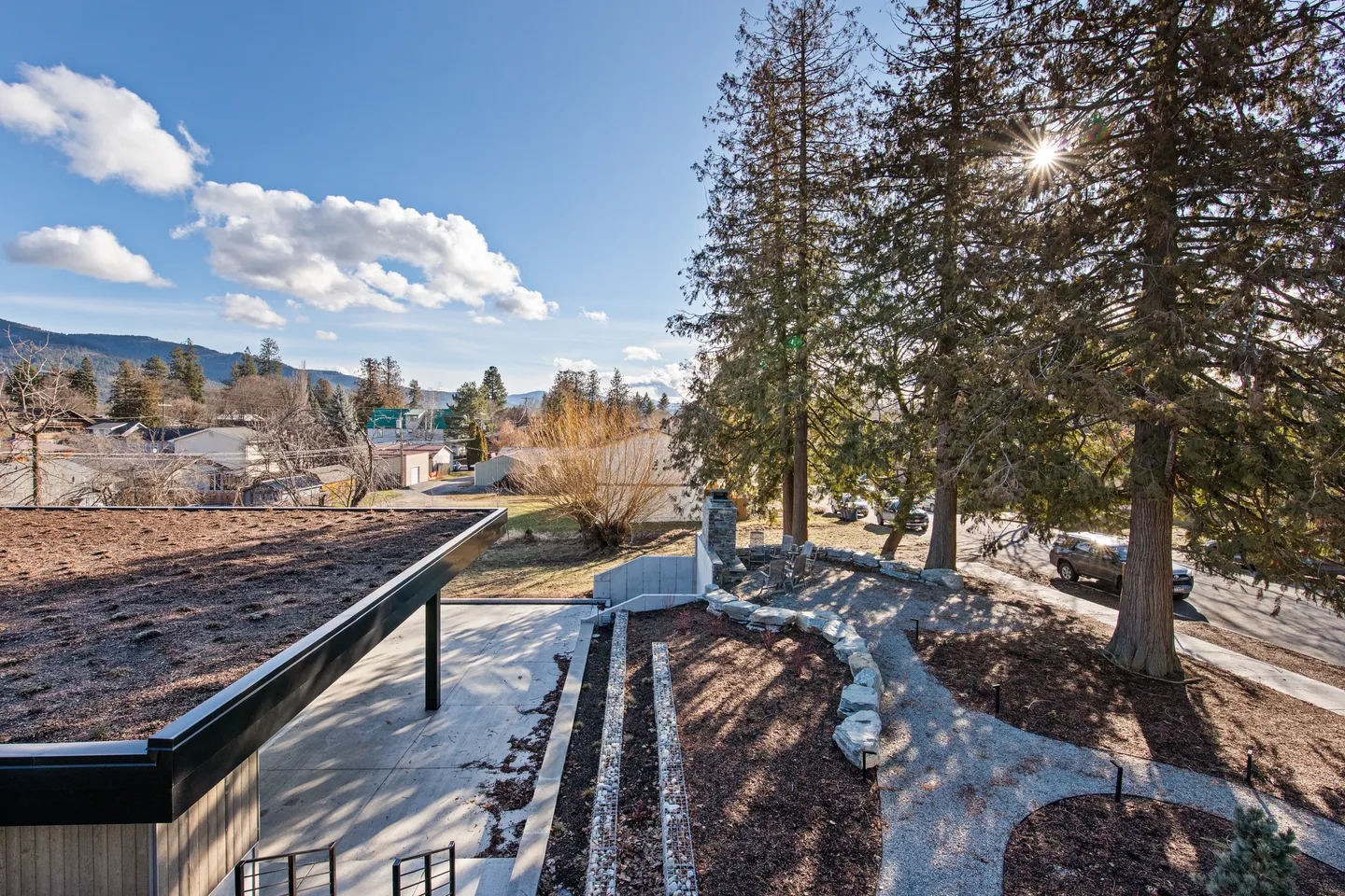 Elevated view of a landscaped outdoor patio, gravel pathways, tall evergreen trees, and distant hills under a sunny sky.