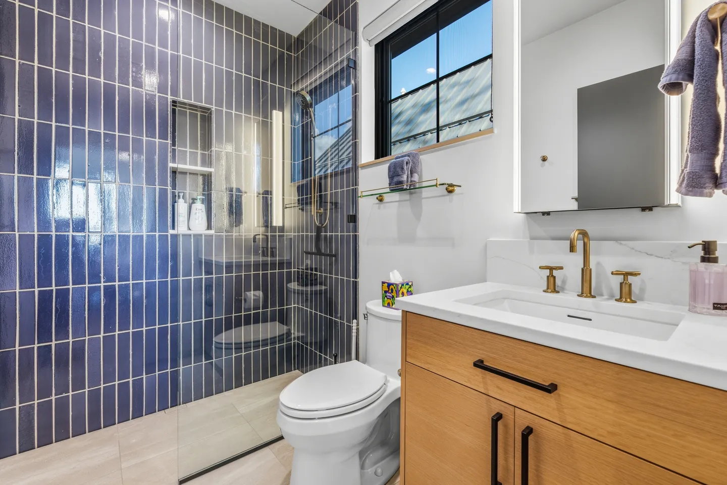 Modern bathroom with blue vertical tile shower, glass door, white toilet, and light wood vanity with brass fixtures.