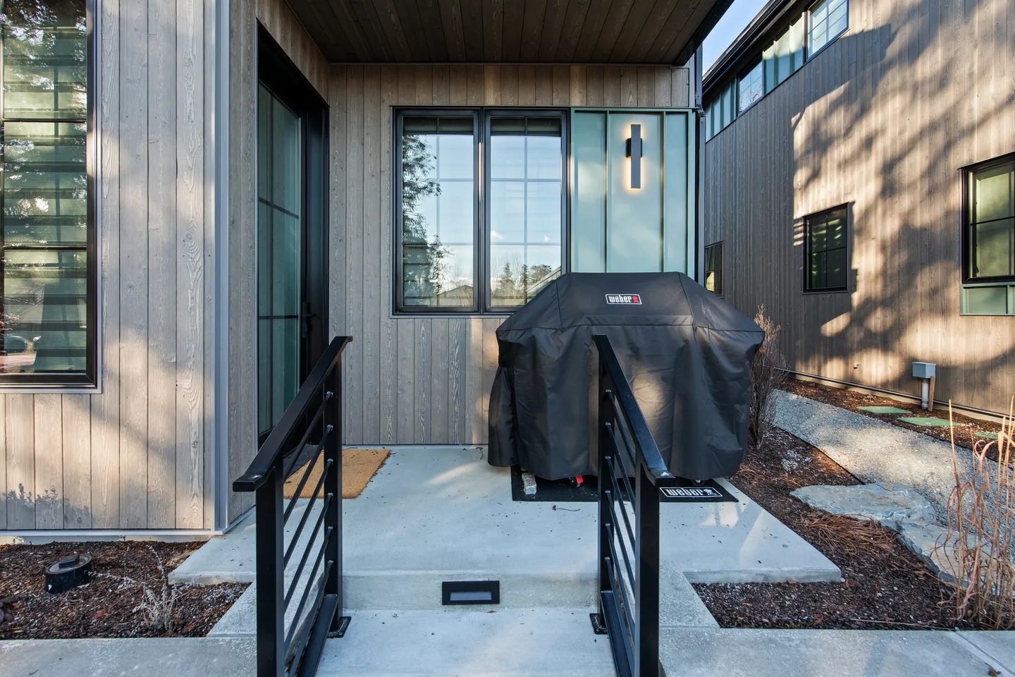 Concrete outdoor porch with black railings, a covered barbecue grill, and vertical wood siding.