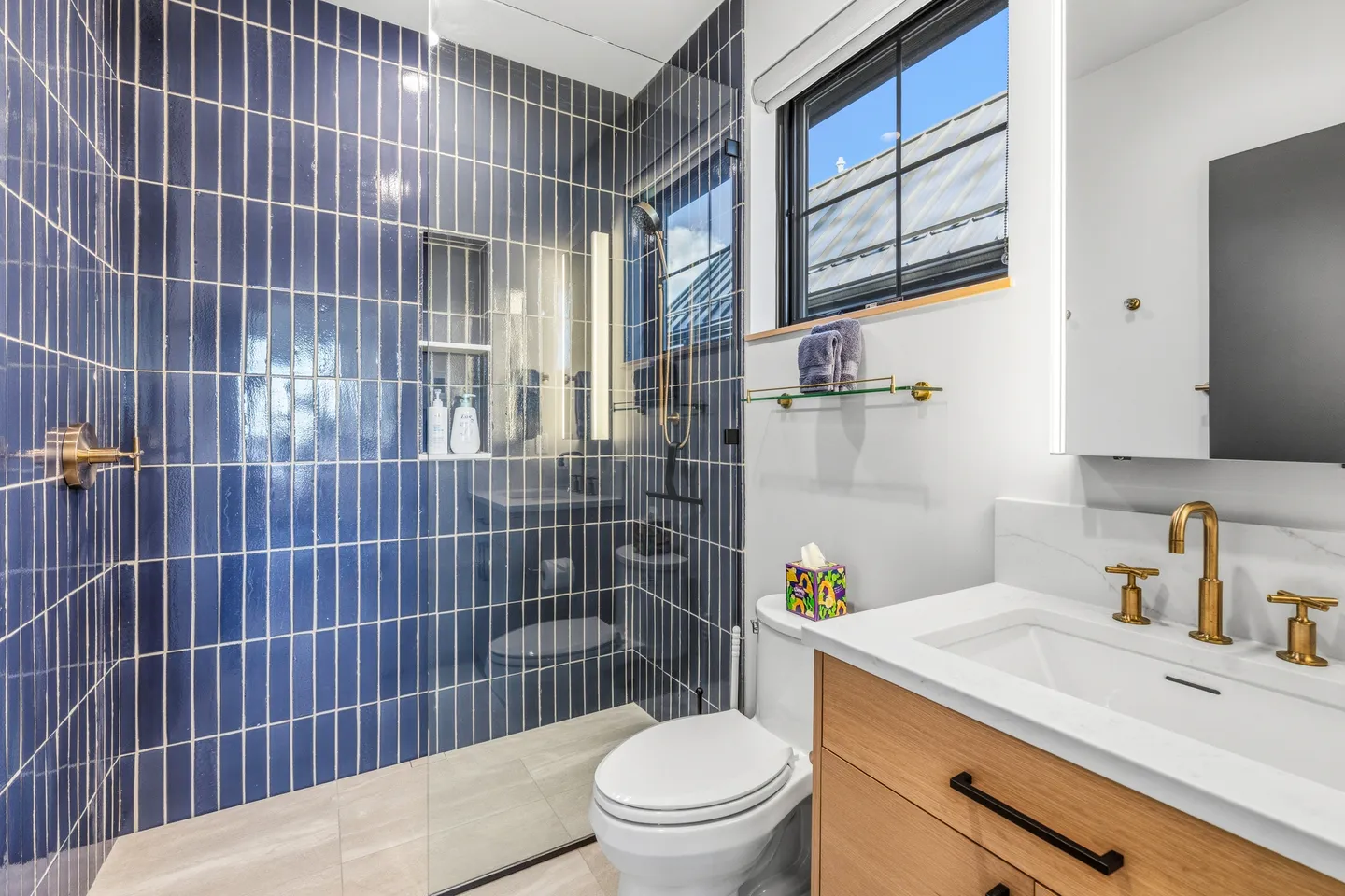 Modern bathroom featuring a walk-in shower with vertical blue tiles, a vanity with gold hardware, and a toilet under a window.