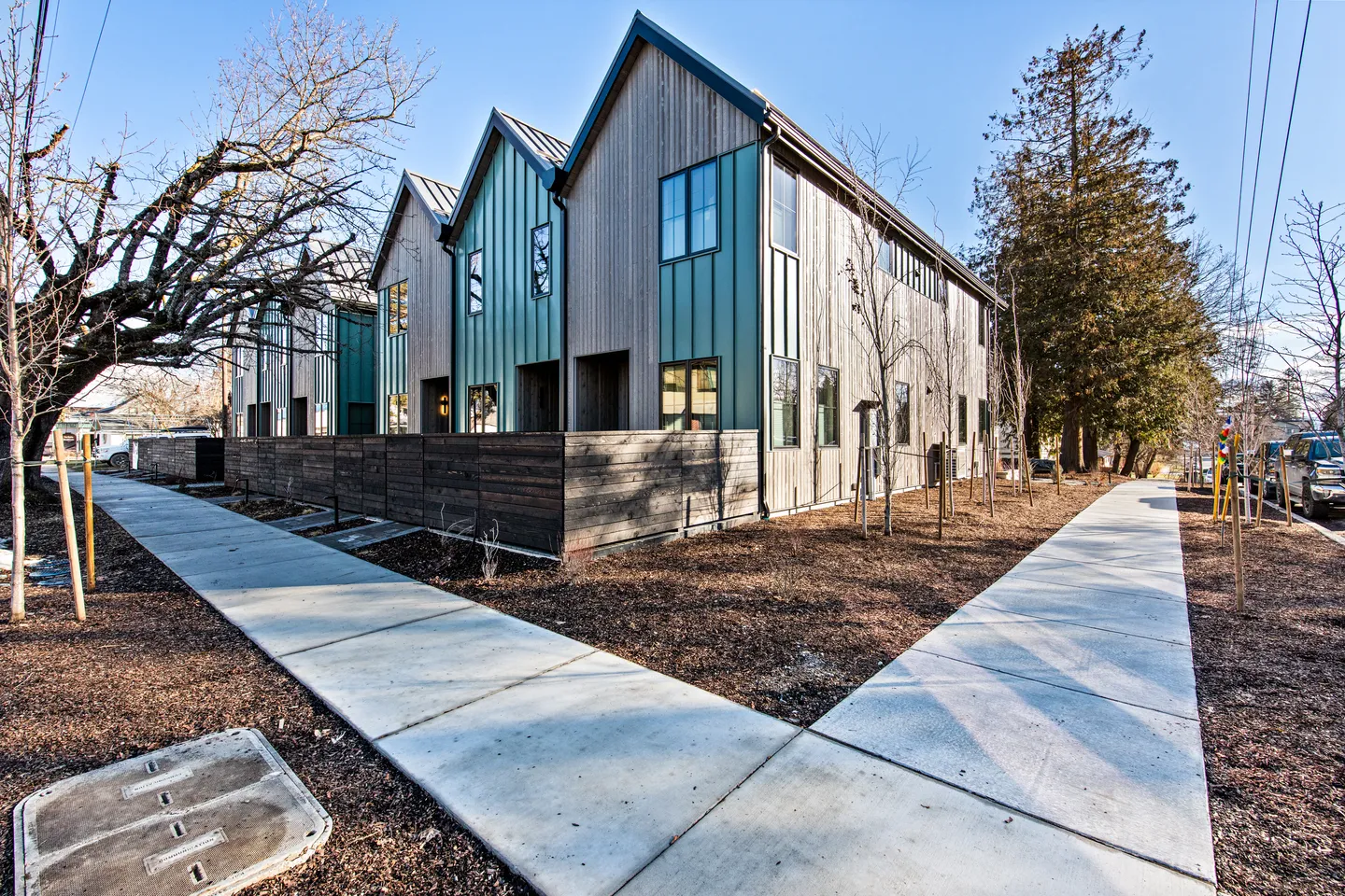 Exterior view of modern two-story townhomes with gabled roofs, vertical wood and teal siding, surrounded by concrete sidewalks and mulch landscaping.