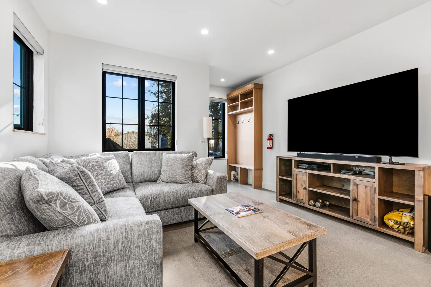 Living room with gray sectional sofa, rustic wooden coffee table, large TV mounted above an entertainment console, and entryway storage bench.