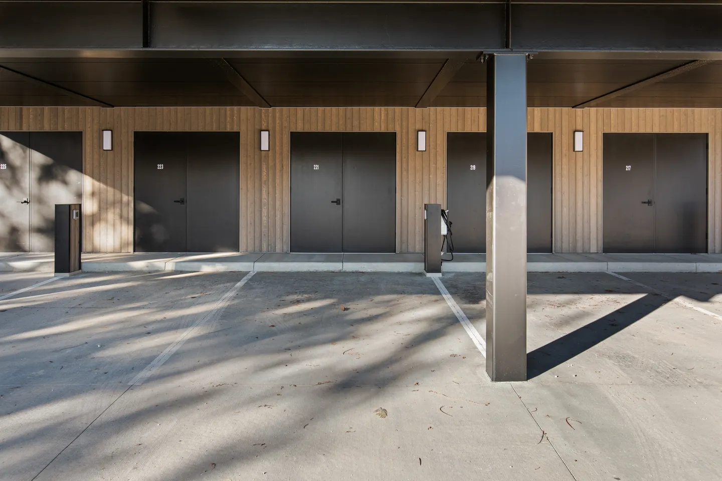 A covered parking area with concrete stalls, EV charging stations, and labeled double doors along a light wood-paneled wall.