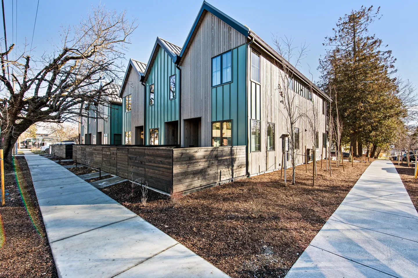Exterior view of modern multi-unit townhomes with gabled roofs, vertical wood siding, and teal panels flanked by concrete walkways and mulch.