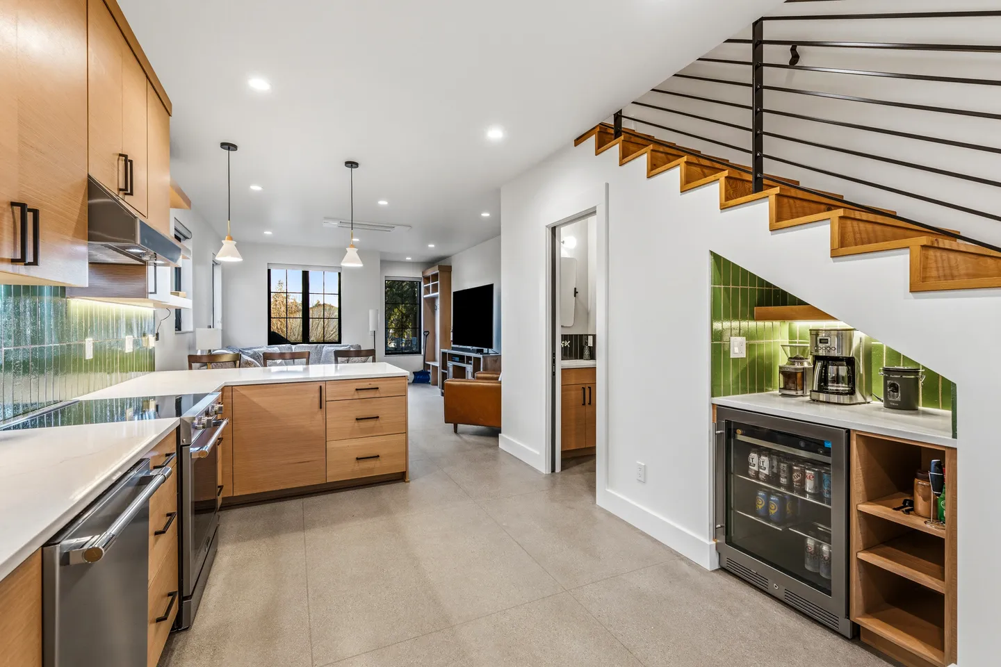 Open-concept modern kitchen and living area with light wood cabinets, green backsplash tile, a staircase with under-stair beverage station, and a view into the living room.
