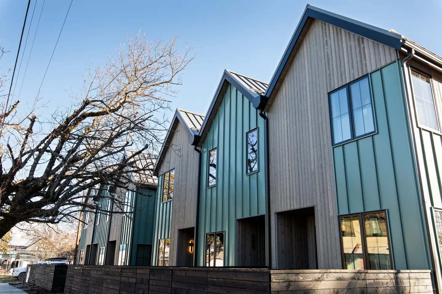 Exterior view of modern townhomes with gabled roofs, vertical teal siding, wood paneling, and a low wooden fence.