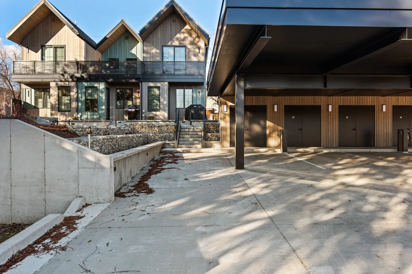 Exterior of modern townhomes with wood siding and black balconies next to a covered carport structure and concrete retaining walls.