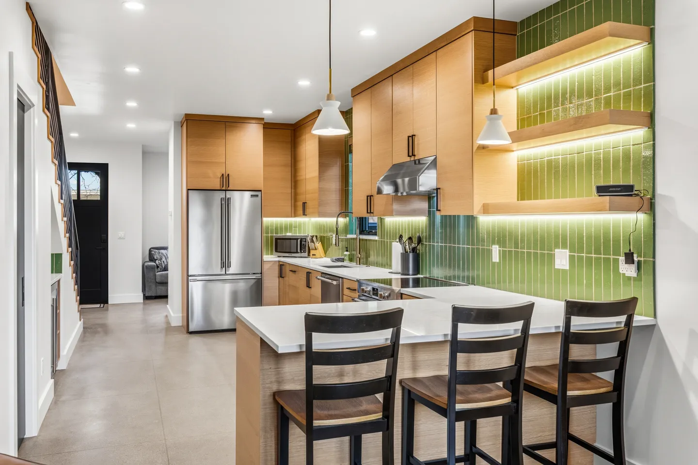 Modern kitchen with light wood cabinetry, green vertical tile backsplash, stainless steel appliances, and a breakfast bar with three stools.