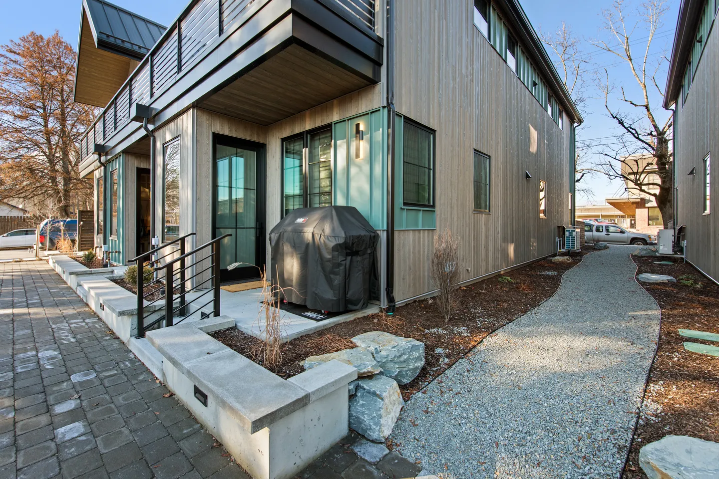Exterior ground-level entrance with a paved walkway, covered grill, and modern wood-sided facade.