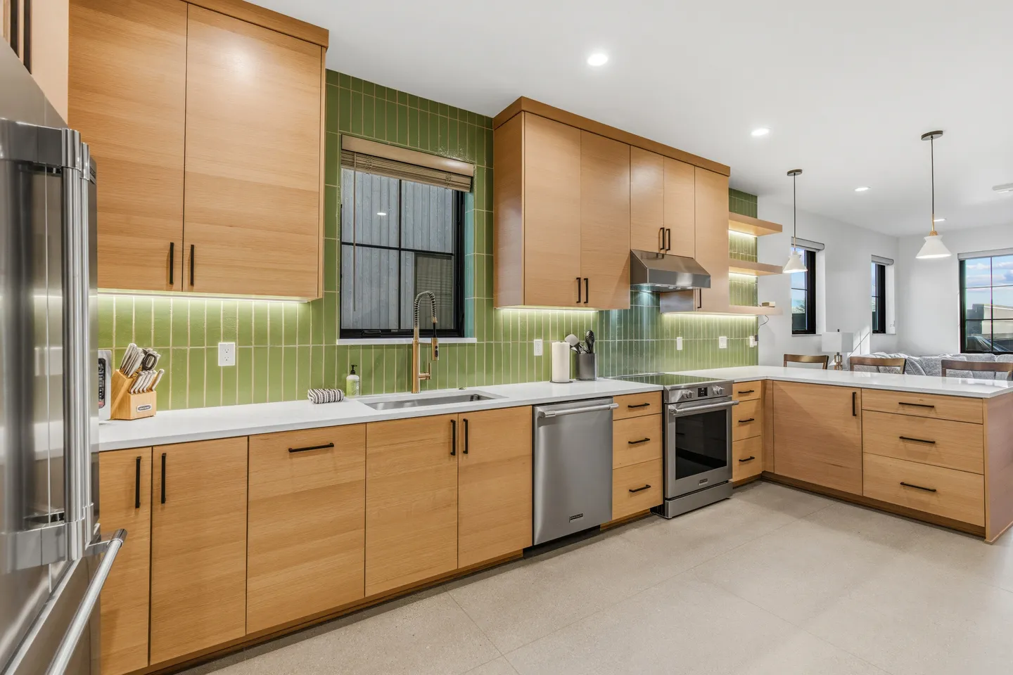 Modern kitchen with light wood cabinets, white countertops, green vertical tile backsplash, and stainless steel appliances.