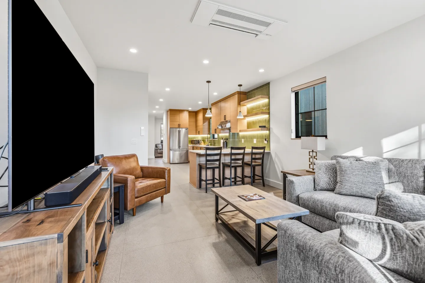 Open-concept living room and kitchen featuring a large TV, leather armchair, gray sofa, coffee table, and an island with barstools.