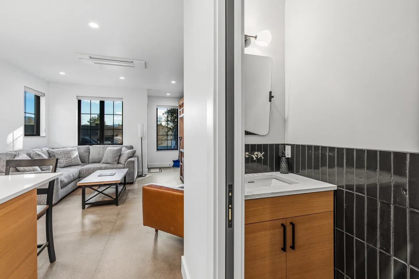A view through an open doorway of a modern bathroom vanity alongside a bright living room with a sectional sofa.