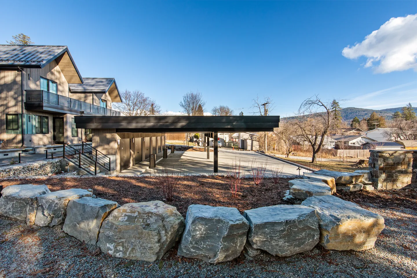 Exterior view of a multi-unit building with a covered carport, stone landscaping border, and mountain views under a clear blue sky.