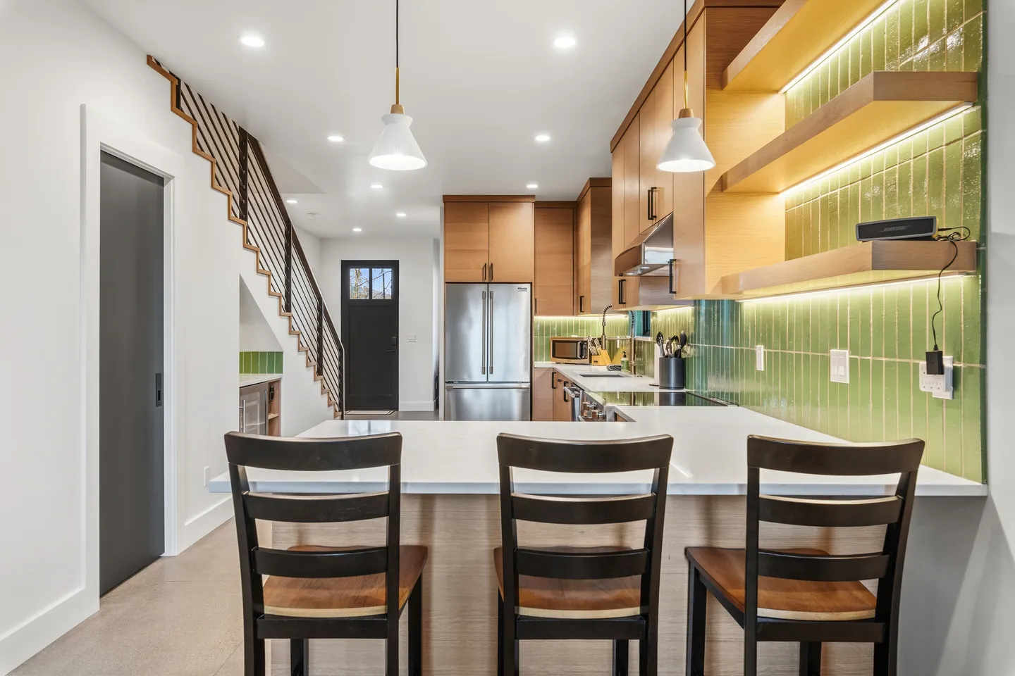 A modern kitchen with wood cabinetry, green tile backsplash, stainless steel appliances, and a breakfast bar with three stools.