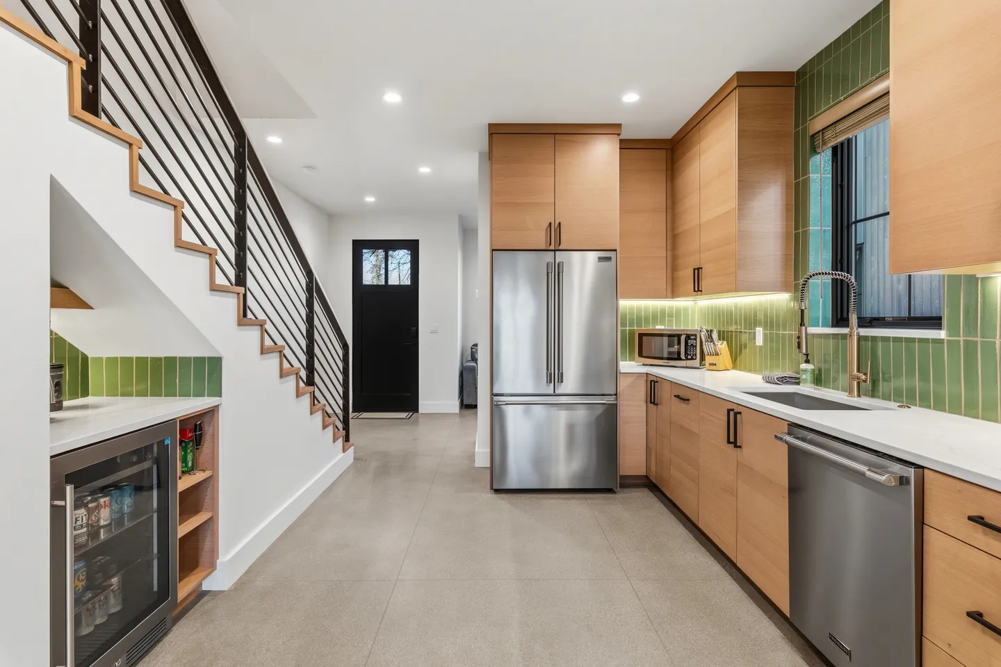 Kitchen with light wood cabinetry, stainless steel appliances, green vertical tile backsplash, and a staircase with a built-in beverage fridge underneath.