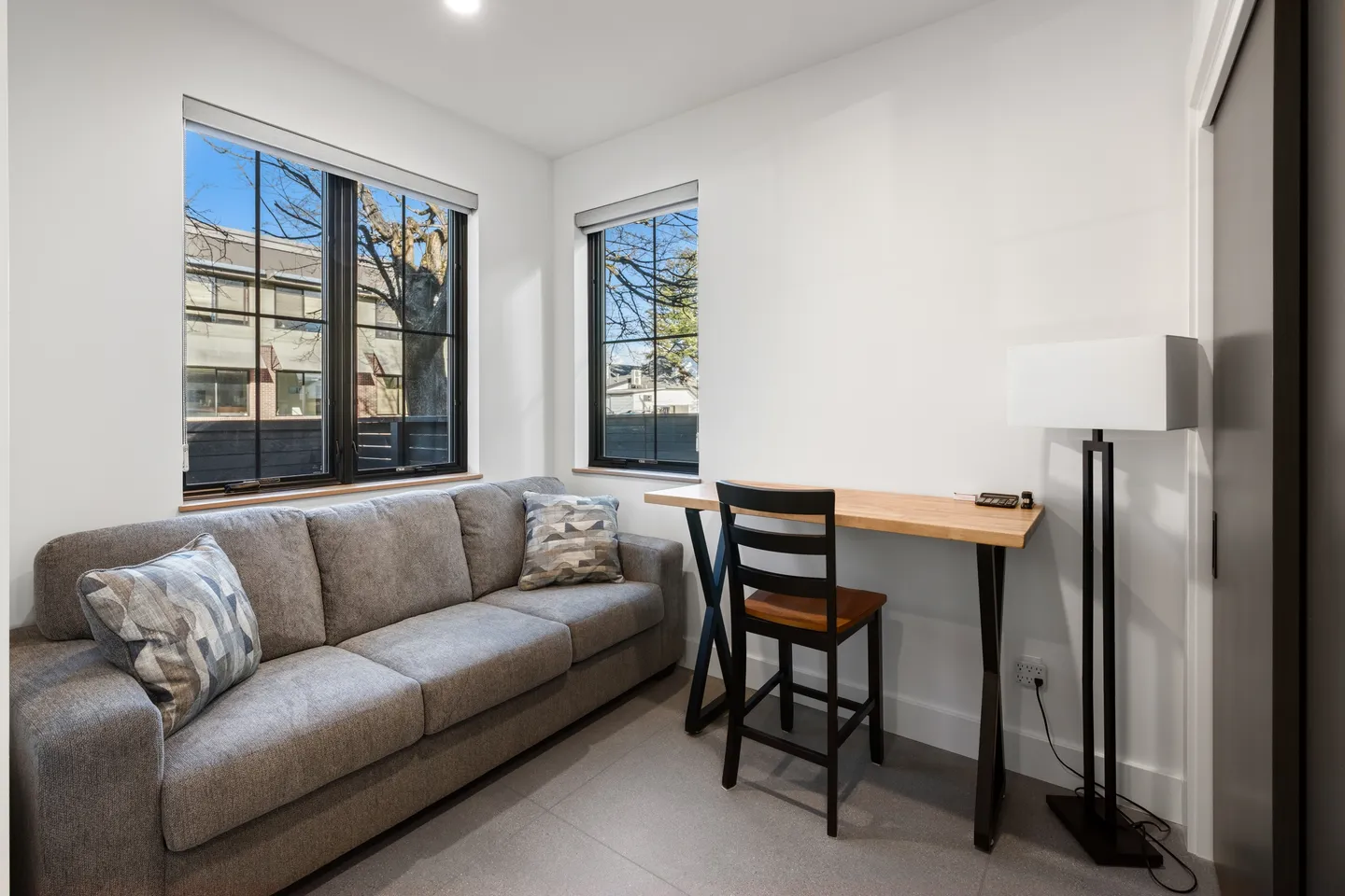 A small room or office nook featuring a gray sofa, a wooden desk with a black chair, and two black-framed windows.