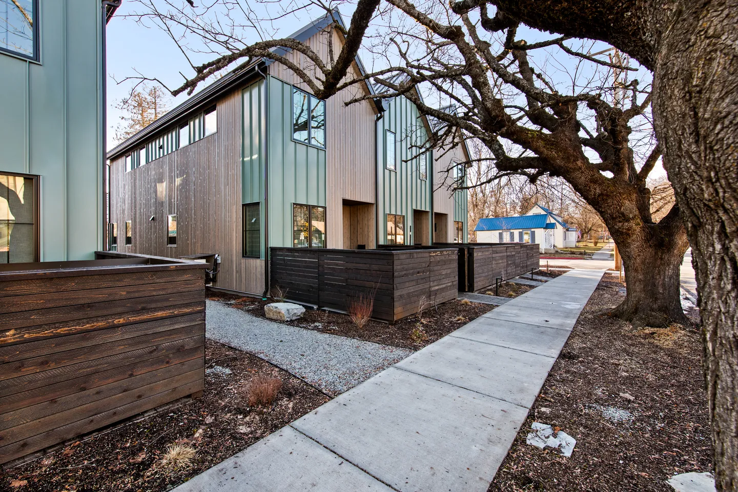 Exterior view of modern townhomes with wood and green metal siding alongside a concrete sidewalk and dark wood fences.